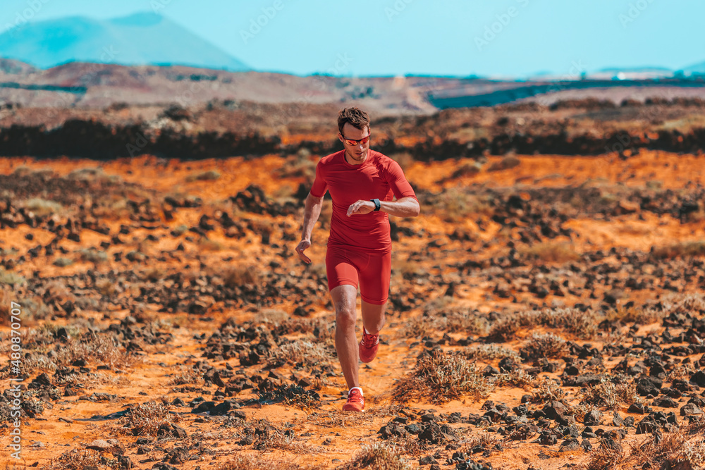 Athlete running checking vitals on smartwatch during endurance race training. Ultra runner man doing triathlon workout on volcanic trail in dramatic red rocks mountain landscape.
