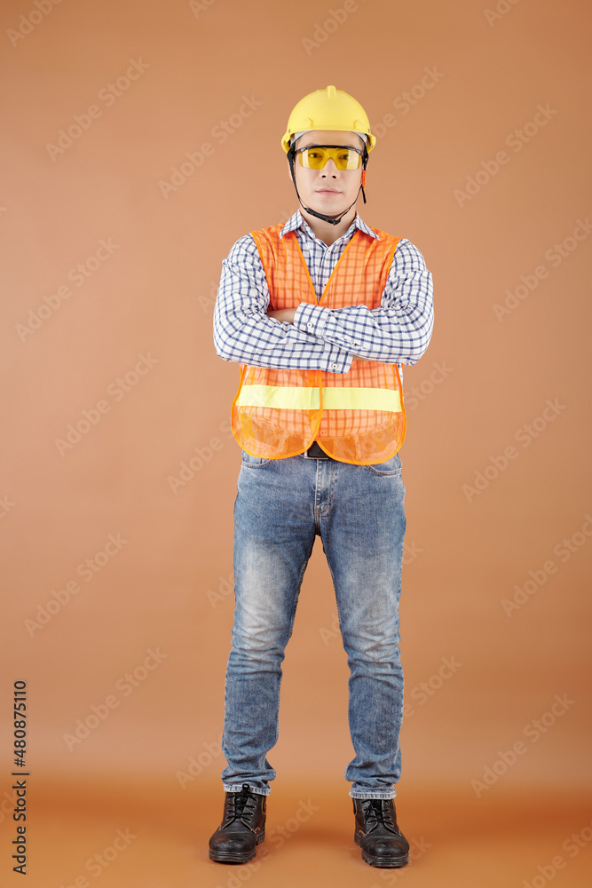 Portrait of civil engineer in orange vest, hard hat and protective glasses crossing arms and looking at camera
