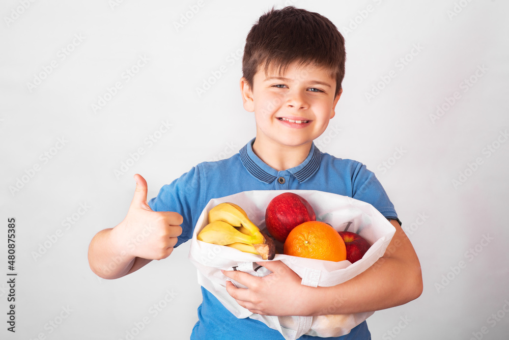 Happy cheerful kid laughs and holds big fabric bag full of healthy fresh fruits and shows sign of approval on white background Happy cheerful kid laughs and holds big fabric bag full of healthy fresh fruits and shows sign of approval on white background