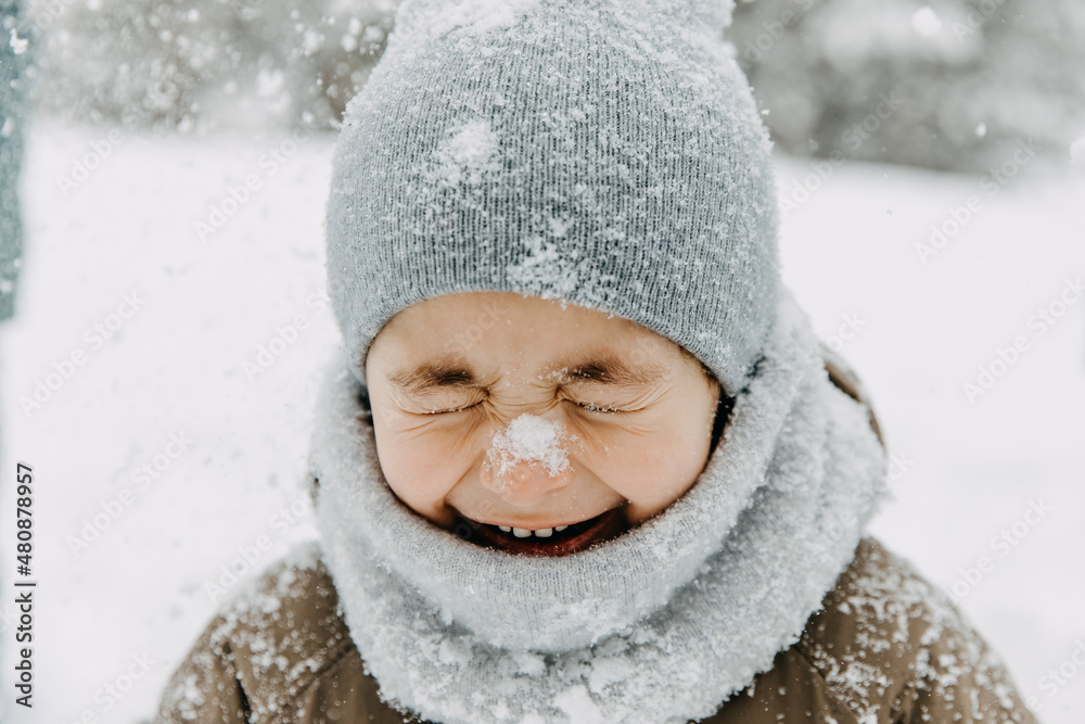Closeup portrait of a little boy with snow on his nose, laughing, with eyes closed, on cold winter day.