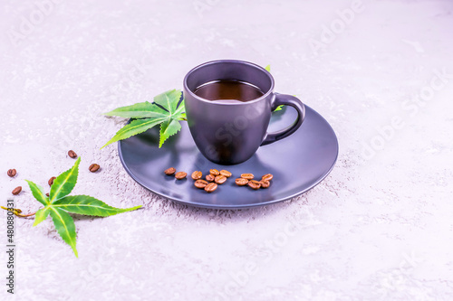 a dark cup with aromatic coffee and connabis leaves on a plate stands on a gray concrete background. The concept of morning coffee for energy.