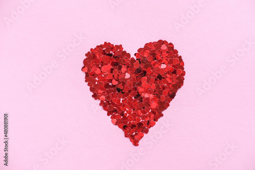 Heart made of shiny red small decorative hearts on a pink background. Flat lay, place for text. Festive minimal composition.