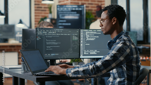 Portrait of african american developer using laptop to write code sitting at desk with multiple screens parsing algorithm in software agency. Coder working on user interface using portable computer. photo