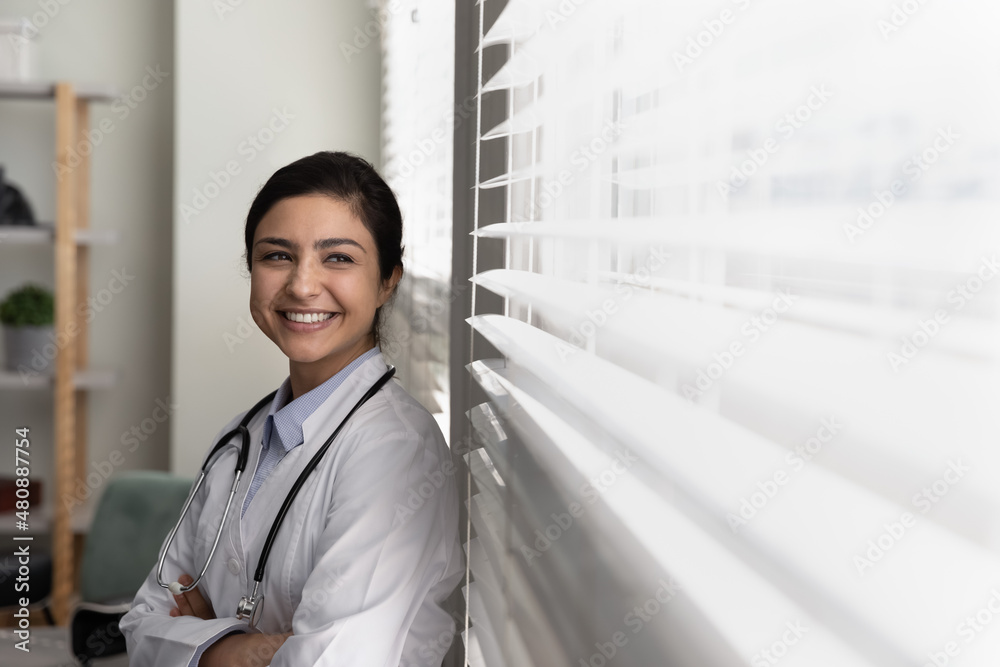 Happy young dreamy successful Indian female general practitioner doctor nurse medical worker looking in distance out of window, visualizing career opportunities or enjoying break time in clinic