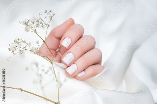 Female hand with white nail design. White nail polish manicure with pink dots. Female hand hold white flower. Woman hand on white fabric background