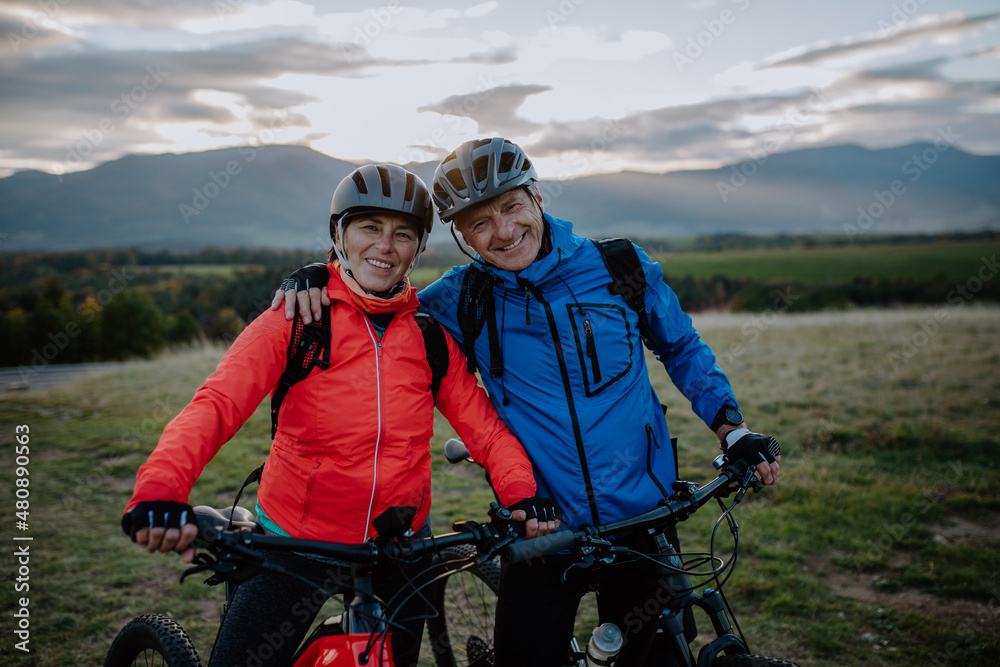 Happy senior couple bikers embracing outdoors in nature in autumn day, looking at camera.