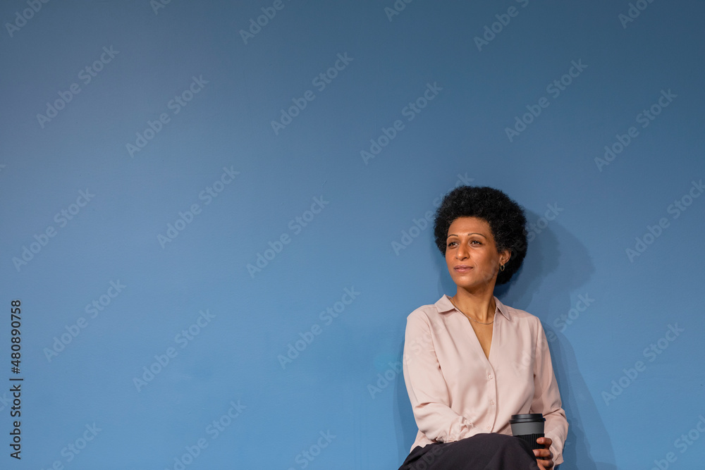 Thoughtful businesswoman with disposable coffee cup in front of blue wall Thoughtful businesswoman with disposable coffee cup in front of blue wall