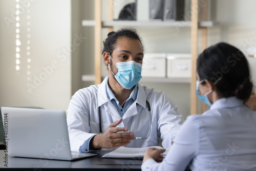 Happy friendly young African American male doctor therapist physician and female Indian patient in facial masks respirators communicating sitting at table in clinic office, medical healthcare concept.