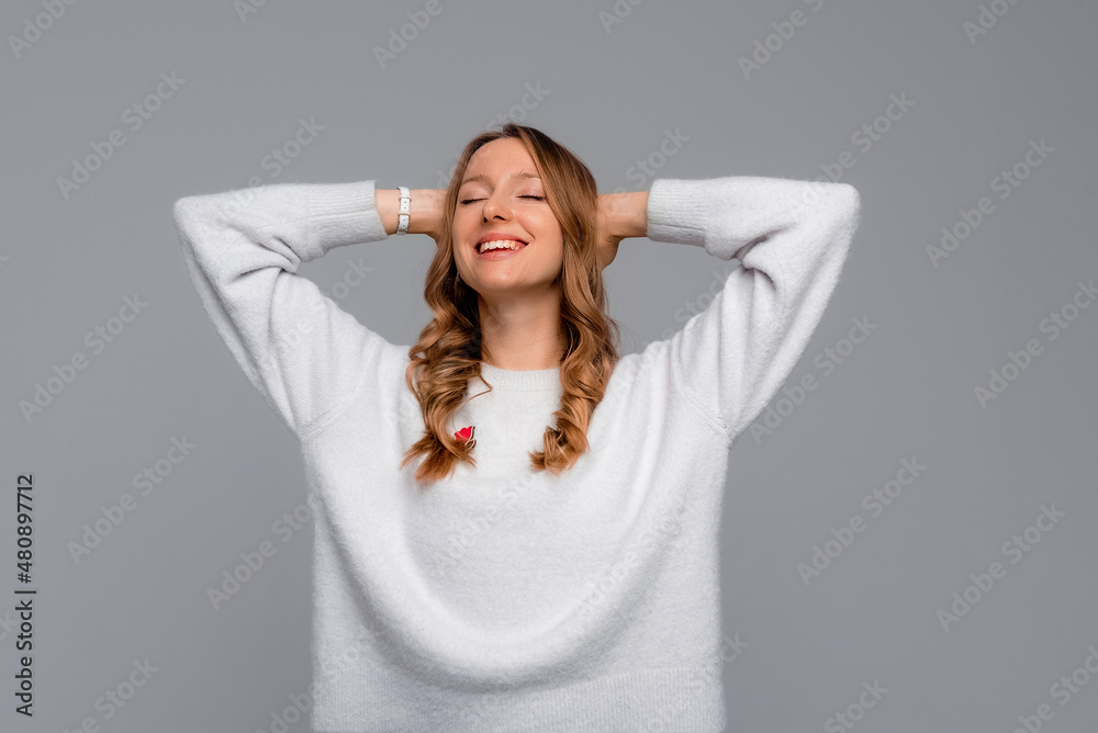 Happy satisfied good-looking young woman. Carefree young woman with a cute grin posing with her hands over her head and eyes closed, standing against gray background Happy satisfied good-looking young woman. Carefree young woman with a cute grin posing with her hands over her head and eyes closed, standing against gray background