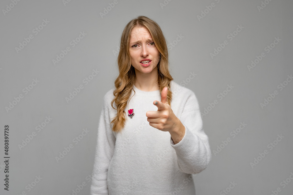 Fototapeta premium Concerned strict young blonde woman pointing index finger on camera choosing you. I blame you in all my problems. Girl wears white sweater, standing against gray background