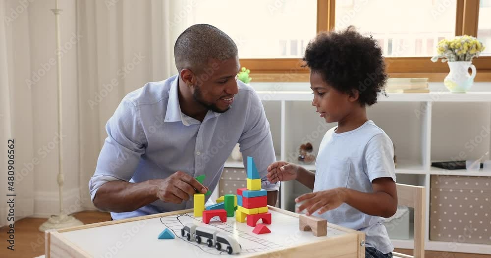 Friendly African American family young father little son sit by table at children room play wooden toy constructor. Grownup elder brother build railway station of colored blocks with younger one child Friendly African American family young father little son sit by table at children room play wooden toy constructor. Grownup elder brother build railway station of colored blocks with younger one child