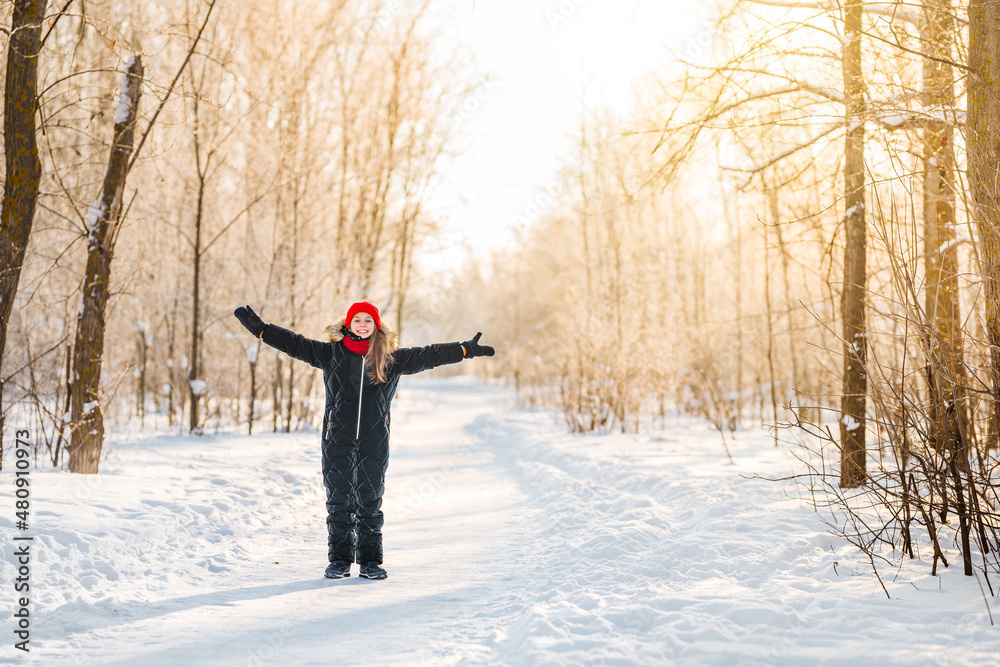 Cute little girl 10 years old in a red hat in a winter forest with sunlight. A little girl is having fun on a winter day. cheerful little girl in a warm jumpsuit