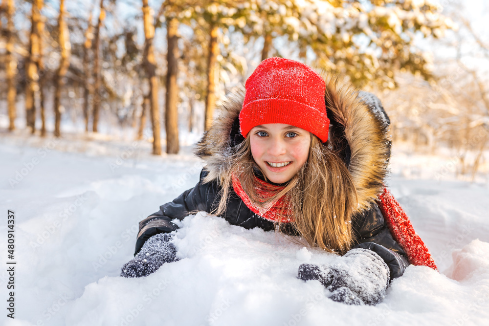 A happy little girl with pink cheeks lies on a snowdrift and laughs in a beautiful winter forest