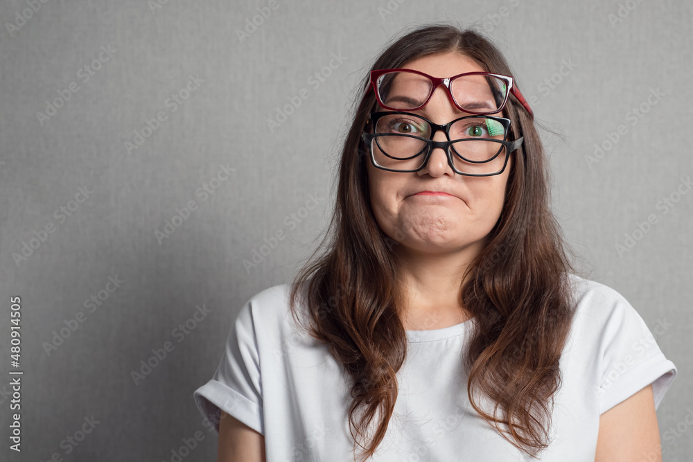 Fototapeta premium Young dark-haired woman with poor eyesight dressed in white t-shirt puts on several pairs of glasses sadly to improve low vision closeup