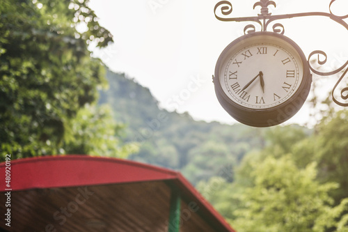 A large clock hangs at the train station in the forest.