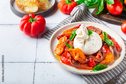 Burrata cheese with baked tomatoes, pepper, red onion and fresh basil on white tile background. Traditional italian food.
