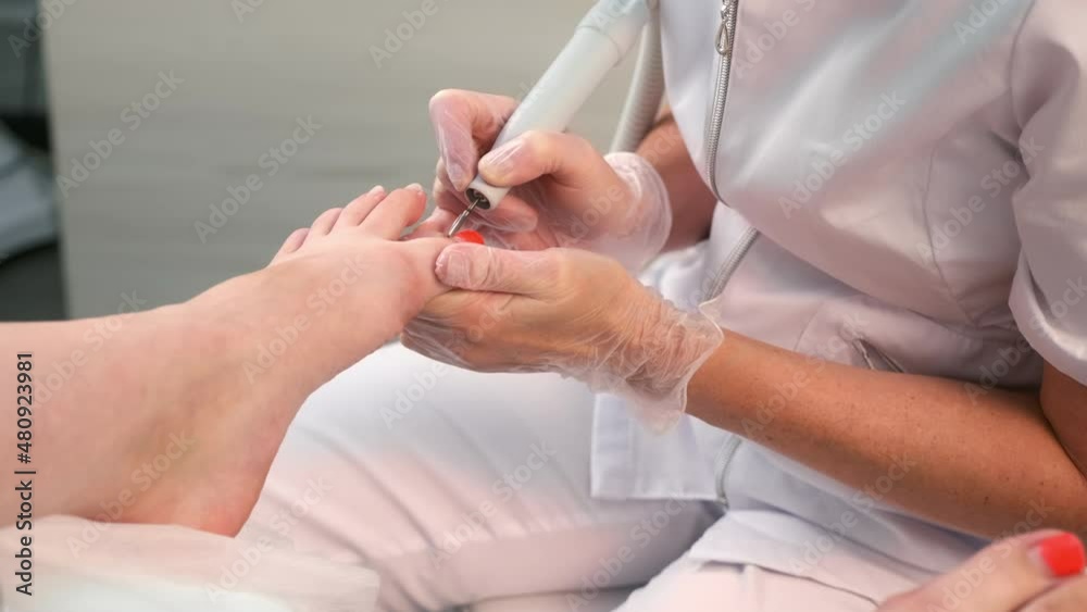 Pedicurist woman removes shellac polish from toes using manicure machine, closeup view. Pedicure Master is removing red gel polish from nails on toes using electric nail drill in cosmetology.