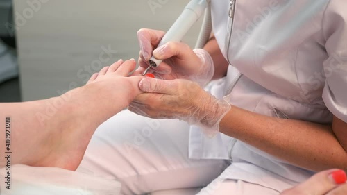 Pedicurist woman removes shellac polish from toes using manicure machine, closeup view. Pedicure Master is removing red gel polish from nails on toes using electric nail drill in cosmetology.