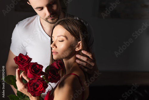 sensual woman with closed eyes holding red roses near man in white t-shirt in dark bedroom.