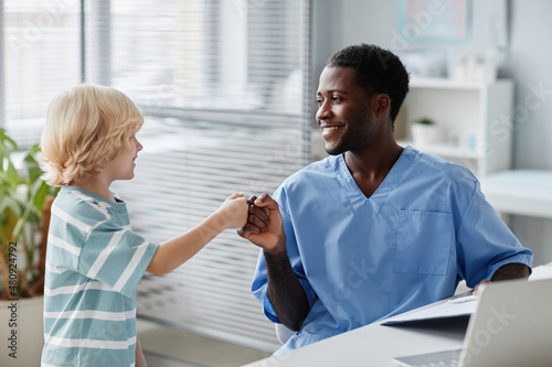 Side view portrait of smiling African-American doctor fist bumping kid during visit to pediatrician in medical clinic