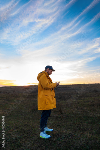 Traveler man in yellow coat with smartphone walking on a hill sunset sky side view. Authentic male tourist with gadget lifestyle. Hipster guy outdoor Solo travel adventure. Active walking backpacking.