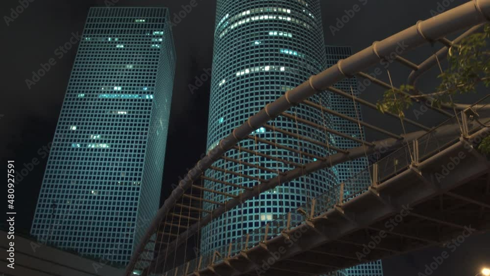 A pedestrian bridge and an ensemble of three high-rise towers against the night sky of Tel Aviv.