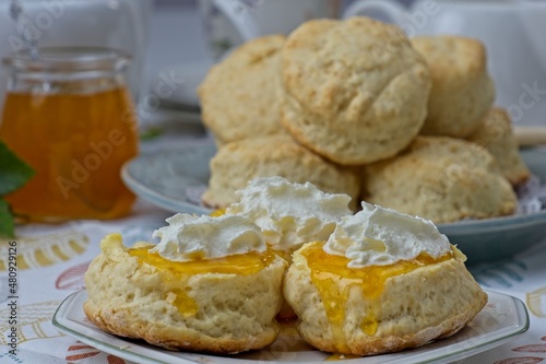 Close up of fresh scones topped with homemade apricot jam and whipped cream with jam jar and scones in background