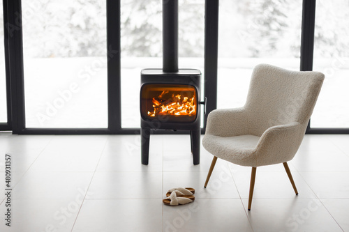 Obraz na plátně Cozy living space by the burning fireplace with chair, cup and slippers on background of snowy landscape