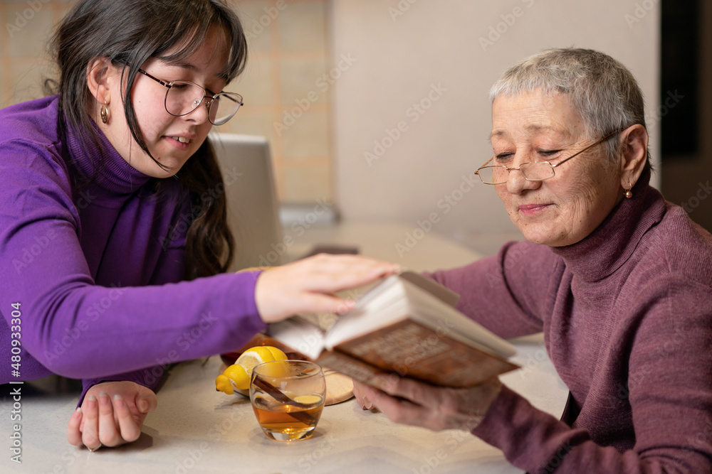 grandmother and granddaughter drink tea sitting at the table and read a book. grandmother and granddaughter drink tea sitting at the table and read a book.