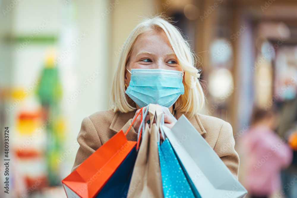 Beautiful girl shopper in medical face mask holding shopping bags in hands. Young happy woman customer in protective mask on face with paper bags. Girl shopaholic. Coronavirus COVID-19, sale, discount