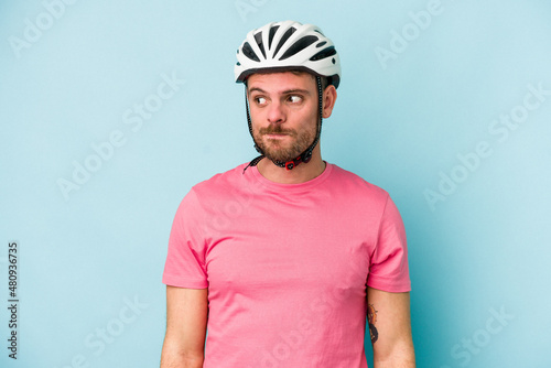 Young caucasian man with bike helmet isolated on blue background confused, feels doubtful and unsure.