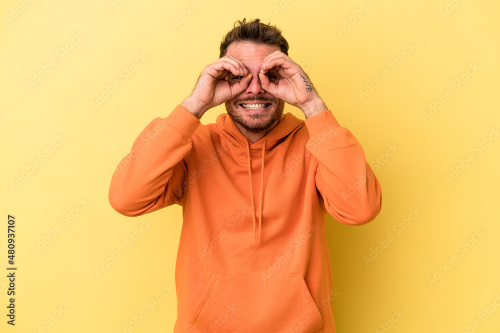 Young caucasian man isolated on yellow background showing okay sign over eyes Young caucasian man isolated on yellow background showing okay sign over eyes