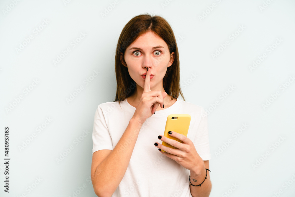 Young English woman holding mobile phone isolated on blue background keeping a secret or asking for silence. Young English woman holding mobile phone isolated on blue background keeping a secret or asking for silence.