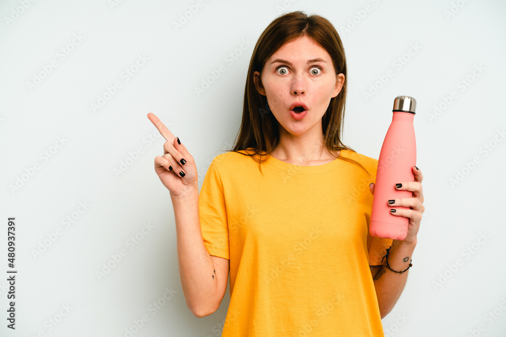 Young English woman holding a thermo isolated on blue background pointing to the side Young English woman holding a thermo isolated on blue background pointing to the side