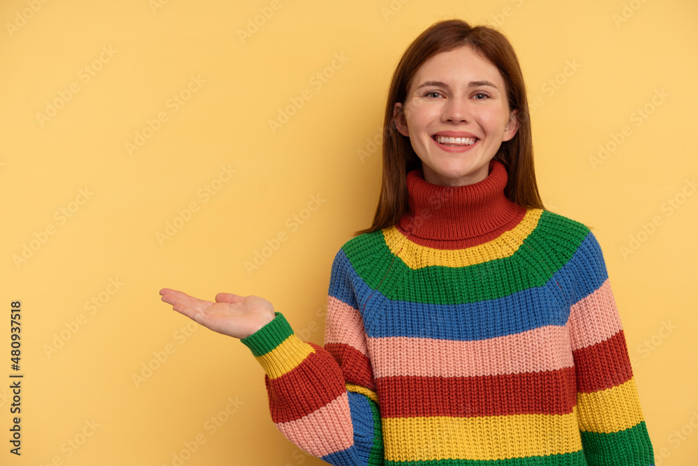 Young English woman isolated on yellow background showing a copy space on a palm and holding another hand on waist. Young English woman isolated on yellow background showing a copy space on a palm and holding another hand on waist.