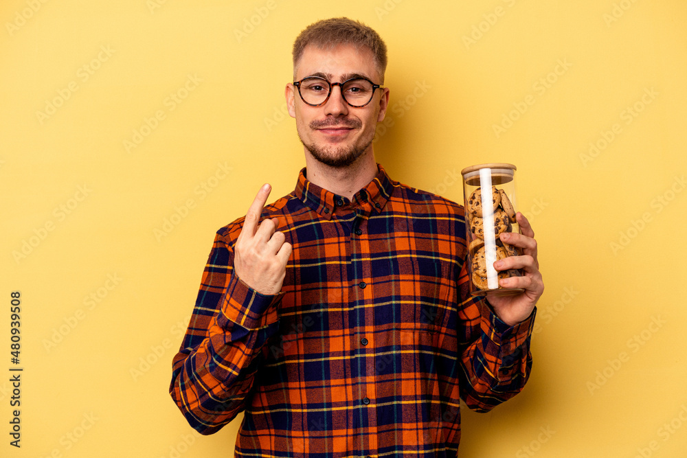 Young caucasian man holding cookies jar isolated on yellow background pointing with finger at you as if inviting come closer. Young caucasian man holding cookies jar isolated on yellow background pointing with finger at you as if inviting come closer.