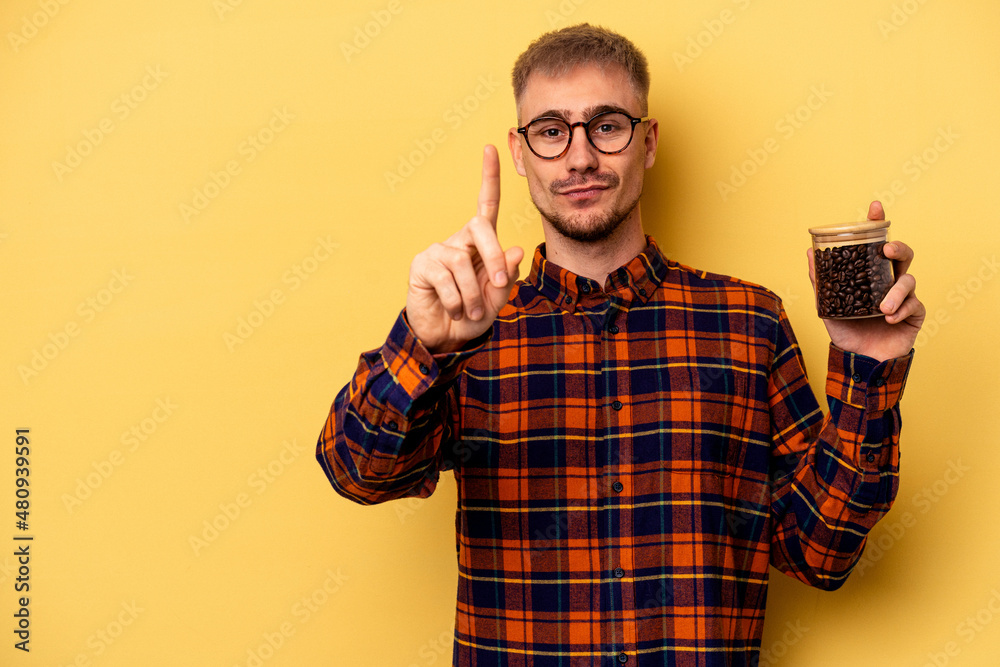 Young caucasian man holding coffee jar isolated on yellow background showing number one with finger.