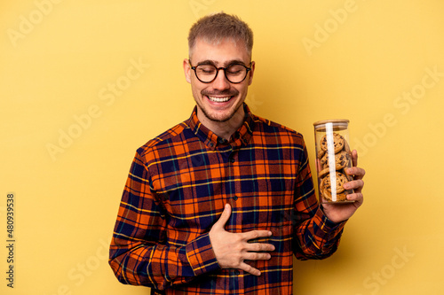 Foto Young caucasian man holding cookies jar isolated on yellow background laughing and having fun