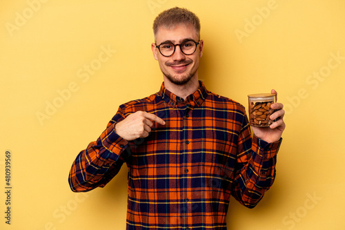 Young caucasian man holding a almond jar isolated on yellow background person pointing by hand to a shirt copy space, proud and confident