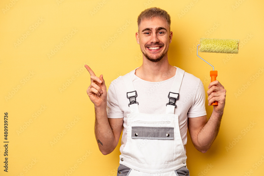 Young painter man isolated on yellow background smiling and pointing aside, showing something at blank space. - obrazy, fototapety, plakaty 