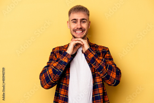 Young caucasian man isolated on yellow background keeps hands under chin, is looking happily aside.