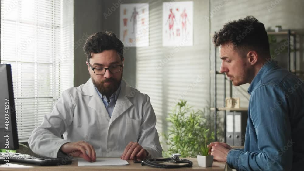 Male Physician Showing Computer to Young Male Patient During Consultation in a Health Clinic. Family Doctor Sitting Behind a Desk in Hospital Office Male Physician Showing Computer to Young Male Patient During Consultation in a Health Clinic. Family Doctor Sitting Behind a Desk in Hospital Office