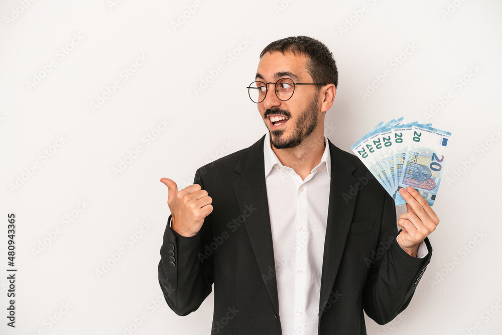 Young caucasian business man holding banknotes isolated on white background points with thumb finger away, laughing and carefree. Young caucasian business man holding banknotes isolated on white background points with thumb finger away, laughing and carefree.