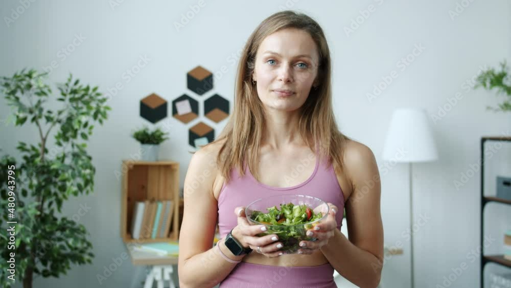 Portrait of pretty blonde holding bowl of salad standing indoors in apartment looking at camera. Young woman is wearing trendy sports clothing. Portrait of pretty blonde holding bowl of salad standing indoors in apartment looking at camera. Young woman is wearing trendy sports clothing.