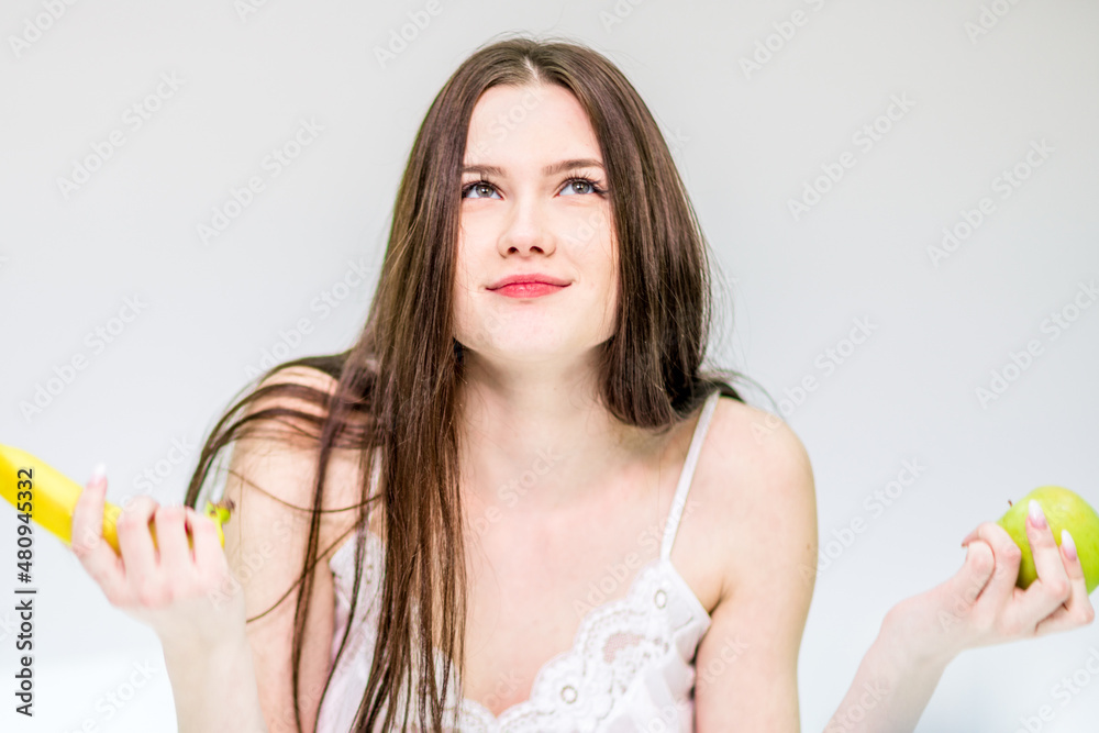 The brunette is having breakfast with fruit. The girl sits on the bed, wrapped in a blanket, and holds a banana and an apple, chooses what to eat The brunette is having breakfast with fruit. The girl sits on the bed, wrapped in a blanket, and holds a banana and an apple, chooses what to eat