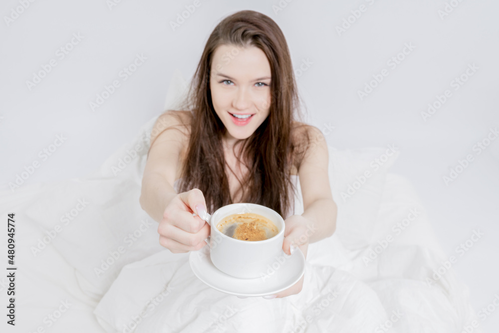 A woman sits in bed and drinks aromatic coffee. Tasty breakfast. The brunette holds a cup of hot drink, offers to try. White bedroom. Good morning A woman sits in bed and drinks aromatic coffee. Tasty breakfast. The brunette holds a cup of hot drink, offers to try. White bedroom. Good morning