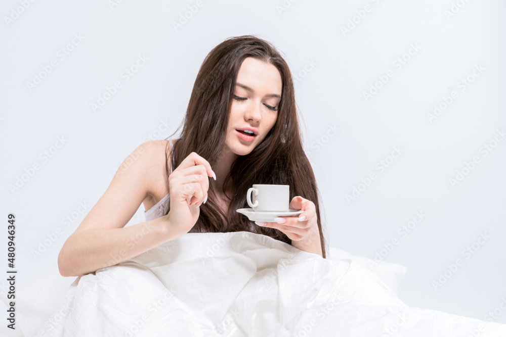 White bedroom. The brunette is holding a cup with a hot drink. A woman is sitting in bed, drinking aromatic coffee. Tasty breakfast. Good morning White bedroom. The brunette is holding a cup with a hot drink. A woman is sitting in bed, drinking aromatic coffee. Tasty breakfast. Good morning