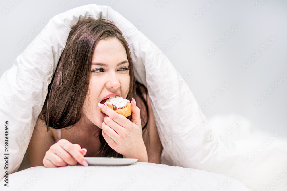 Calories. A cute woman with appetite eats a cupcake lying on the bed under the covers. Dessert. Breakfast in bed Calories. A cute woman with appetite eats a cupcake lying on the bed under the covers. Dessert. Breakfast in bed