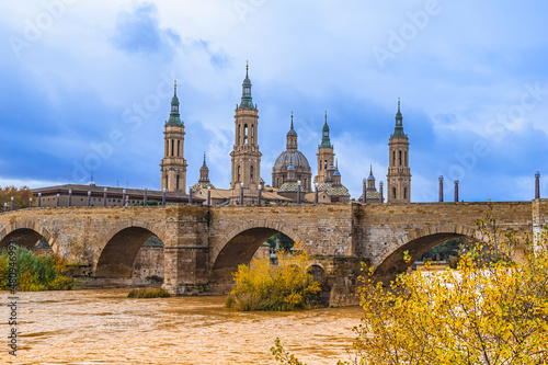 Ancient stone Puente de Piedra Bridge over Ebro River with yellow water on the backdrop of the towers of Cathedral-Basilica of Our Lady of the Pillar in Saragossa, Spain. Autumn medieval cityscape