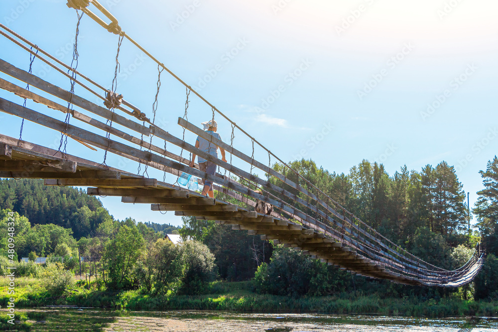 Fototapeta premium Woman with dog walks along old wooden hanging bridge on river in summer sunny day. Natural landscape. Dog on bridge